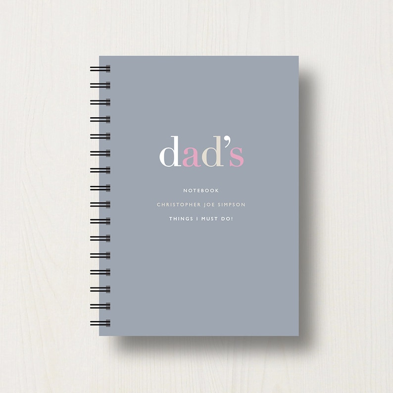 May include: A gray notebook with a spiral binding. The cover has the text "dad's" in white and pink letters. Below the text, it says "Notebook", "Christopher Joe Simpson", and "Things I Must Do!"