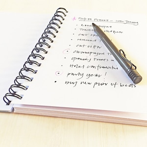 May include: A white notebook with a black spiral binding. The notebook is open to a page with a handwritten to-do list for a trip to France. The list includes items such as "renew passport", "travel insurance", "confirm hotel", and "buy new pair of boots".