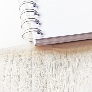May include: A white spiral bound notebook with a silver binding. The notebook is lying open on a light wood surface.