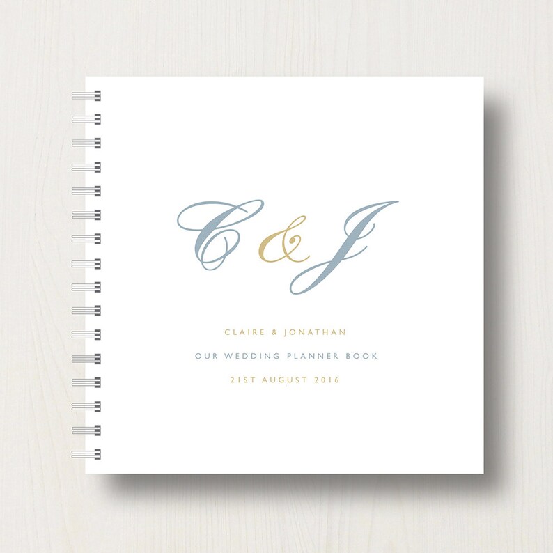 May include: A white wedding planner book with a blue and gold C&J monogram. The text "Claire & Jonathan Our Wedding Planner Book 21st August 2016" is printed below the monogram.