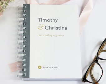 Può includere: Un planner di nozze bianco a spirale con il testo "Timothy & Christina our wedding organiser" e "27th July 2020" sulla copertina.