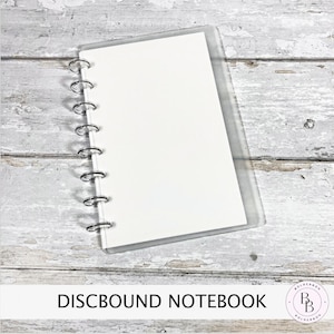 May include: A discbound notebook with a clear plastic cover and silver metal rings. The notebook has white paper inside. The words "DISCBOUND NOTEBOOK" are printed in black at the bottom of the image.