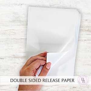 May include: A stack of white double-sided release paper. The paper is being held by a hand, with one sheet partially peeled back to show the adhesive side.