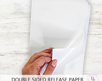 May include: A stack of white double-sided release paper. The paper is being held by a hand, with one sheet partially peeled back to show the adhesive side.