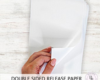 Może przedstawiać: Stos białego papieru do odklejania dwustronnego. Papier jest trzymany przez rękę, a jeden arkusz jest częściowo odklejony, aby pokazać klejącą stronę.