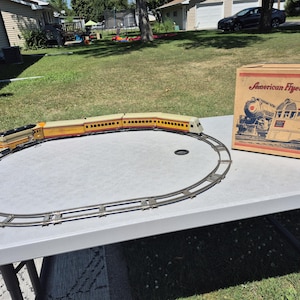 May include: A vintage American Flyer Trains toy set featuring a yellow and red locomotive with matching passenger cars on a circular metal track. The set includes a cardboard box with the text American Flyer Trains and a graphic illustration.