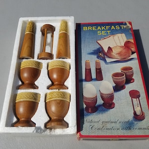 May include: A wooden breakfast set in a white box. The set includes four egg cups, two salt and pepper shakers, and an egg timer. The box has the words "Breakfast Set" printed on it. The egg cups and shakers are made of natural grained wood with ceramic accents.