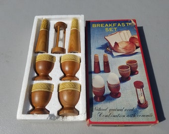 May include: A wooden breakfast set in a white box. The set includes four egg cups, two salt and pepper shakers, and an egg timer. The box has the words "Breakfast Set" printed on it. The egg cups and shakers are made of natural grained wood with ceramic accents.