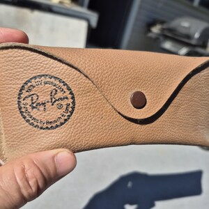 May include: Tan leather sunglasses case with a snap closure. The case features the Ray-Ban logo and text indicating 100% UV protection. The case is designed to protect sunglasses and has a textured surface.