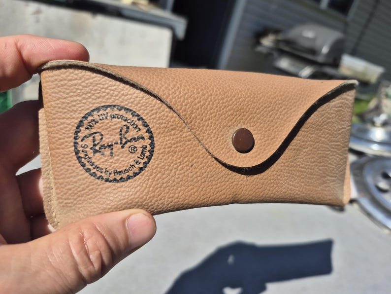 May include: Tan leather sunglasses case with a snap closure. The case features the Ray-Ban logo and text indicating 100% UV protection. The case is designed to protect sunglasses and has a textured surface.