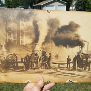 May include: A black and white photograph of three vintage fire engines with smoke billowing from their stacks. The fire engines are lined up in a row, and there are people standing around them. The fire engines are in front of a building with windows. The photograph is faded and has a brown tint.