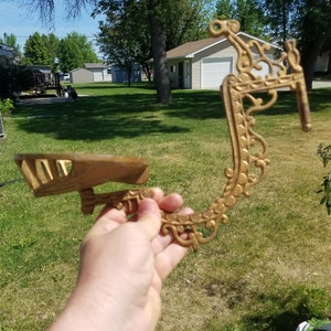 May include: Ornate, gold-colored metal bracket with a decorative, scrollwork design. The bracket has a small shelf and a curved, openwork frame. The piece is held up against a green grassy background.