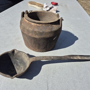May include: A rustic, dark brown cast iron pot with two handles and a matching ladle. The ladle has a long handle and a rounded bowl. The pot and ladle appear to be vintage or antique.