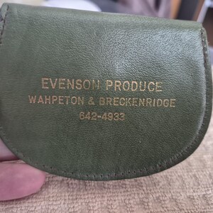 May include: A small, olive green leather coin purse with a curved top. The text "EVENSON PRODUCE" is embossed in gold lettering, along with the locations "WAHPETON & BRECKENRIDGE" and a phone number. The purse is held by a hand.