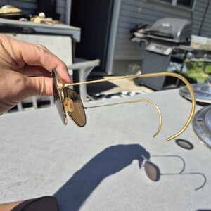 May include: Gold-framed sunglasses with brown lenses. The sunglasses have a classic aviator style with a curved bridge and temples. The lenses are a warm brown color, and the frames are a shiny gold. The sunglasses are held up.