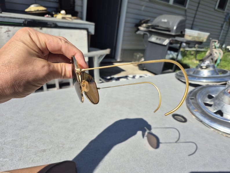 May include: Gold-framed sunglasses with brown lenses. The sunglasses have a classic aviator style with a curved bridge and temples. The lenses are a warm brown color, and the frames are a shiny gold. The sunglasses are held up.