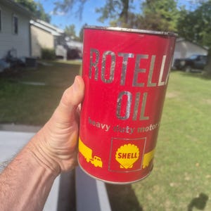 May include: A cylindrical red metal motor oil can featuring the silver text ROTELLA OIL and the words heavy duty motor oil. The can displays a yellow Shell logo inside a square border and yellow graphic illustrations of trucks on the lower section.