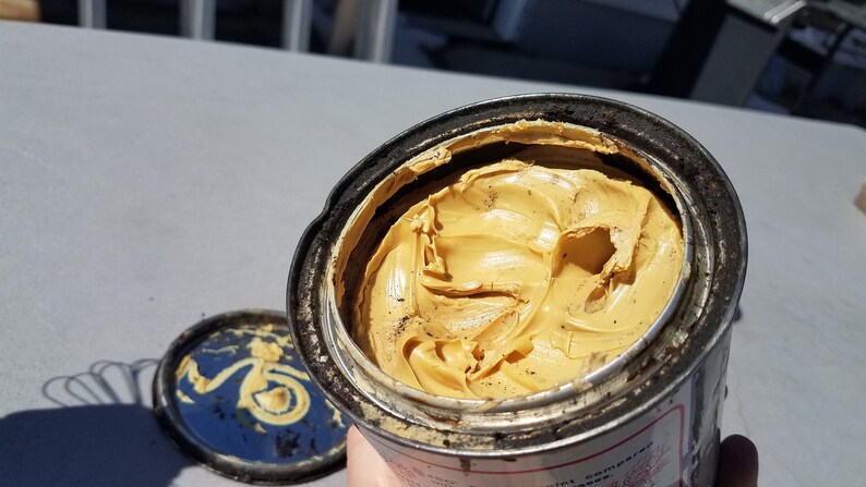 May include: A partially full tin of yellow grease. The tin is dirty and has some rust on it.