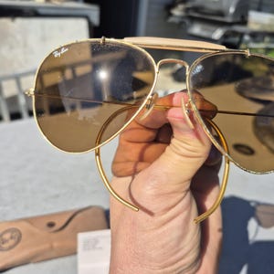 May include: Gold-framed sunglasses with brown lenses. The sunglasses have a double bridge and a leather brow bar. The Ray-Ban logo is visible on the lens. A brown case and paperwork are in the background.
