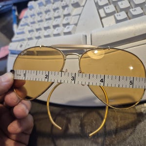 May include: Gold-framed aviator sunglasses with amber lenses. The glasses are being measured with a tape measure, showing a width of approximately 5 inches. The classic design features a double bridge and curved temples.