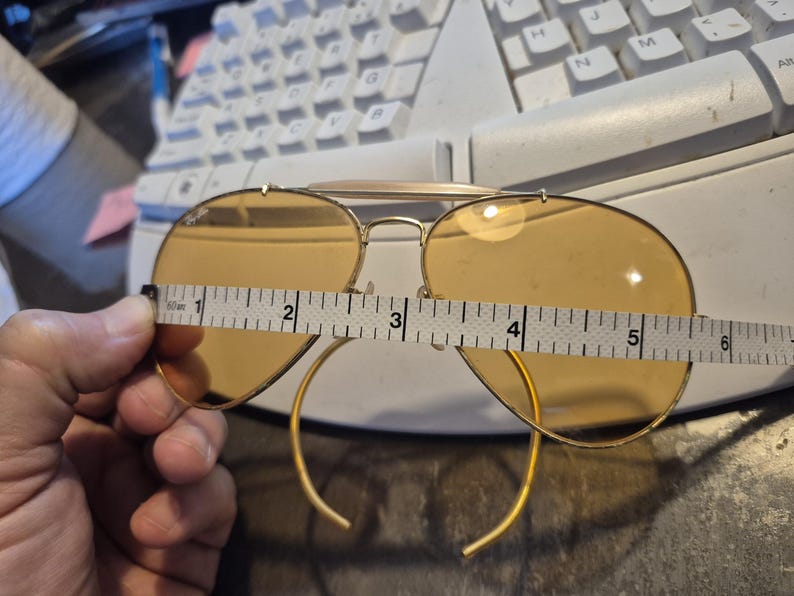 May include: Gold-framed aviator sunglasses with amber lenses. The glasses are being measured with a tape measure, showing a width of approximately 5 inches. The classic design features a double bridge and curved temples.
