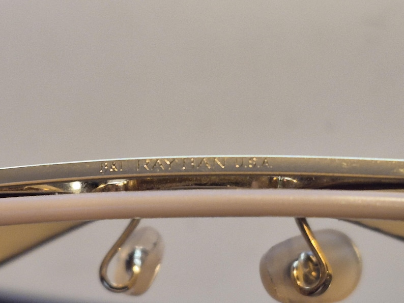 May include: Close-up of the top frame of Ray-Ban sunglasses. The gold-colored frame has the text "PAT. RAY-BAN U.S.A." etched into it. The image also shows the nose pads and part of the lenses.