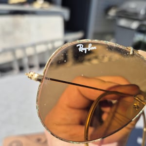 May include: Close-up of Ray-Ban sunglasses with gold frames and brown lenses. The Ray-Ban logo is visible on the lens. The sunglasses have a classic aviator style. The lenses are slightly reflective.