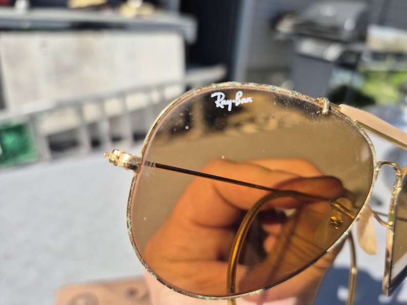 May include: Close-up of Ray-Ban sunglasses with gold frames and brown lenses. The Ray-Ban logo is visible on the lens. The sunglasses have a classic aviator style. The lenses are slightly reflective.