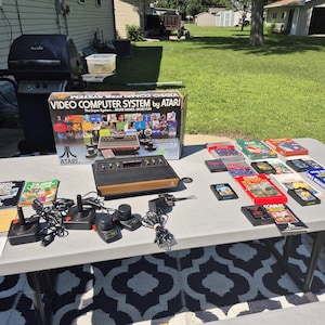 May include: An Atari Video Computer System box set with a console, controllers, and game cartridges on a table. The box reads "Video Computer System by Atari". The games include titles like "Space Invaders" and "Combat".