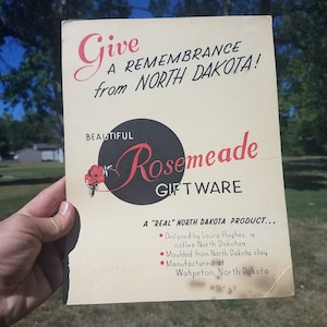 May include: A vintage advertisement for Rosemeade Giftware, featuring text in red and black. The text reads "Give a Remembrance from North Dakota!" and "Beautiful Rosemeade Giftware." The ad is held in front of a green grassy background.