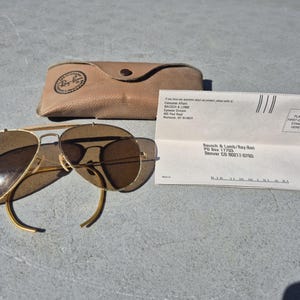 May include: Vintage Ray-Ban sunglasses with a gold frame and brown lenses, alongside a tan leather case and a folded paper with text. The sunglasses have a classic aviator design. The case has a Ray-Ban logo.