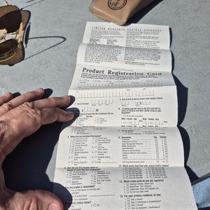 May include: A pair of gold-framed sunglasses with brown lenses rests near a folded product registration card. The card, with the text "Product Registration Card," is partially filled out. A tan glasses case is also visible.