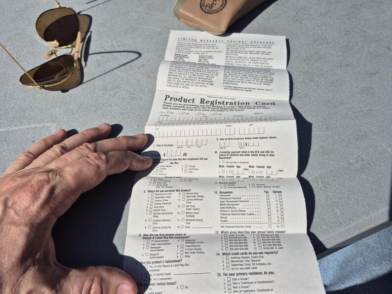 May include: A pair of gold-framed sunglasses with brown lenses rests near a folded product registration card. The card, with the text "Product Registration Card," is partially filled out. A tan glasses case is also visible.