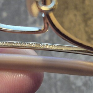 May include: Close-up of a pair of gold-toned Ray-Ban sunglasses. The text "B&L RAY-BAN 62 [] 14" is etched on the temple arm. The sunglasses have a classic design, with a metal frame and tinted lenses. The image highlights the brand's logo and model details.