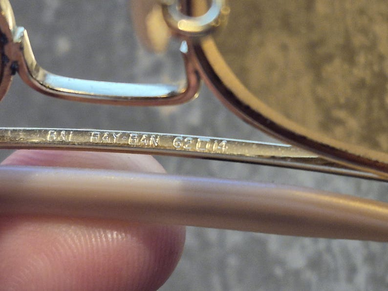 May include: Close-up of a pair of gold-toned Ray-Ban sunglasses. The text "B&L RAY-BAN 62 [] 14" is etched on the temple arm. The sunglasses have a classic design, with a metal frame and tinted lenses. The image highlights the brand's logo and model details.