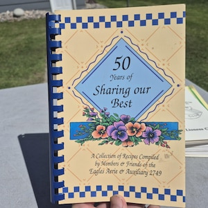 May include: A spiral-bound cookbook with a cream-colored cover featuring the text "50 Years of Sharing our Best." The cover has a blue diamond shape with the title, floral accents, and the text "A Collection of Recipes Compiled by Members & Friends of the Eagles Aerie & Auxiliary 2749."