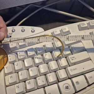 May include: A pair of round, gold-rimmed glasses with a curved temple arm, measured with a ruler. The glasses are resting on a white computer keyboard. The ruler shows the glasses are approximately 2 inches in length.