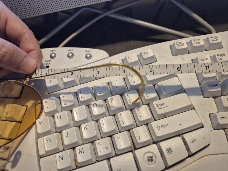 May include: A pair of round, gold-rimmed glasses with a curved temple arm, measured with a ruler. The glasses are resting on a white computer keyboard. The ruler shows the glasses are approximately 2 inches in length.