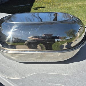 May include: A shiny, oval-shaped, stainless steel roasting pan with a lid and two handles. The lid reflects the surrounding environment. The pan is sitting on a light gray surface.