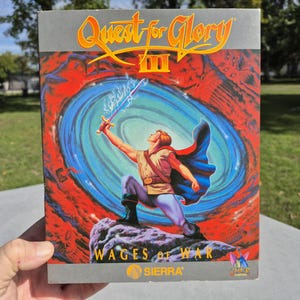 May include: A rectangular video game box for Quest for Glory III: Wages of War by Sierra. The cover art features a character in a tan tunic and blue cape standing on a rock, holding a glowing sword toward a swirling blue portal against a red background.
