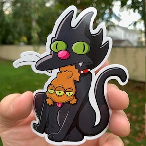 May include: A black cat with green eyes and a pink nose sits with a three-eyed orange alien creature on its back. The cat is wearing a red collar with a silver buckle.