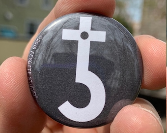 May include: A black and white button with a stylized symbol of a cross with a circle on top and a curved line underneath. The text "2019 FESETTE STUDIO" is printed around the edge of the button.