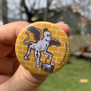 May include: A yellow button with a brick pattern background. A winged skeleton monkey with a top hat is holding a purse with a skeleton animal inside. The button says "©2019 Fesette Studio".