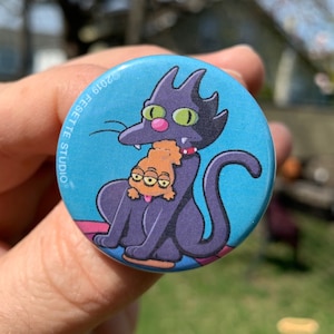 May include: A blue button with a black cat holding a three-eyed orange cat. The button has the text "STUDIO FESETE 2016" on the top.