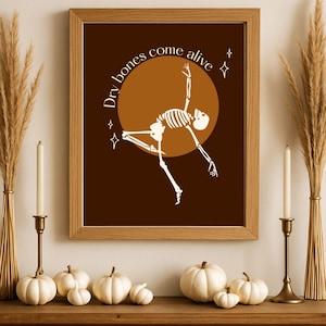 May include: A framed print with the text "Dry bones come alive" and a skeleton illustration. The artwork is set against a brown background with a circular design. The image is part of a Halloween or fall-themed display, with pumpkins, candles, and decorative wheat stalks.