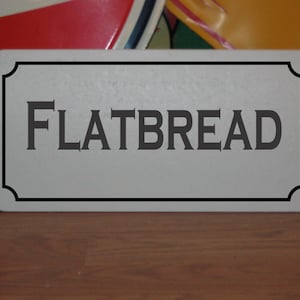 May include: A white rectangular sign with a black border and the word "FLATBREAD" printed in black.