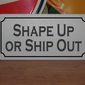May include: A rectangular metal sign with the words "SHAPE UP OR SHIP OUT" in dark gray lettering. The sign has a light gray background and a black border. The sign is on a wooden surface.