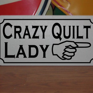 May include: A white sign with black text and a black pointing hand. The sign reads "Crazy Quilt Lady".