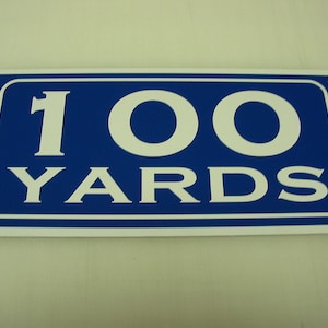 May include: A blue and white sign with the text "100 YARDS" in white lettering.