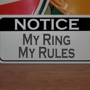 May include: A silver metal sign with black lettering that reads "NOTICE MY RING MY RULES".